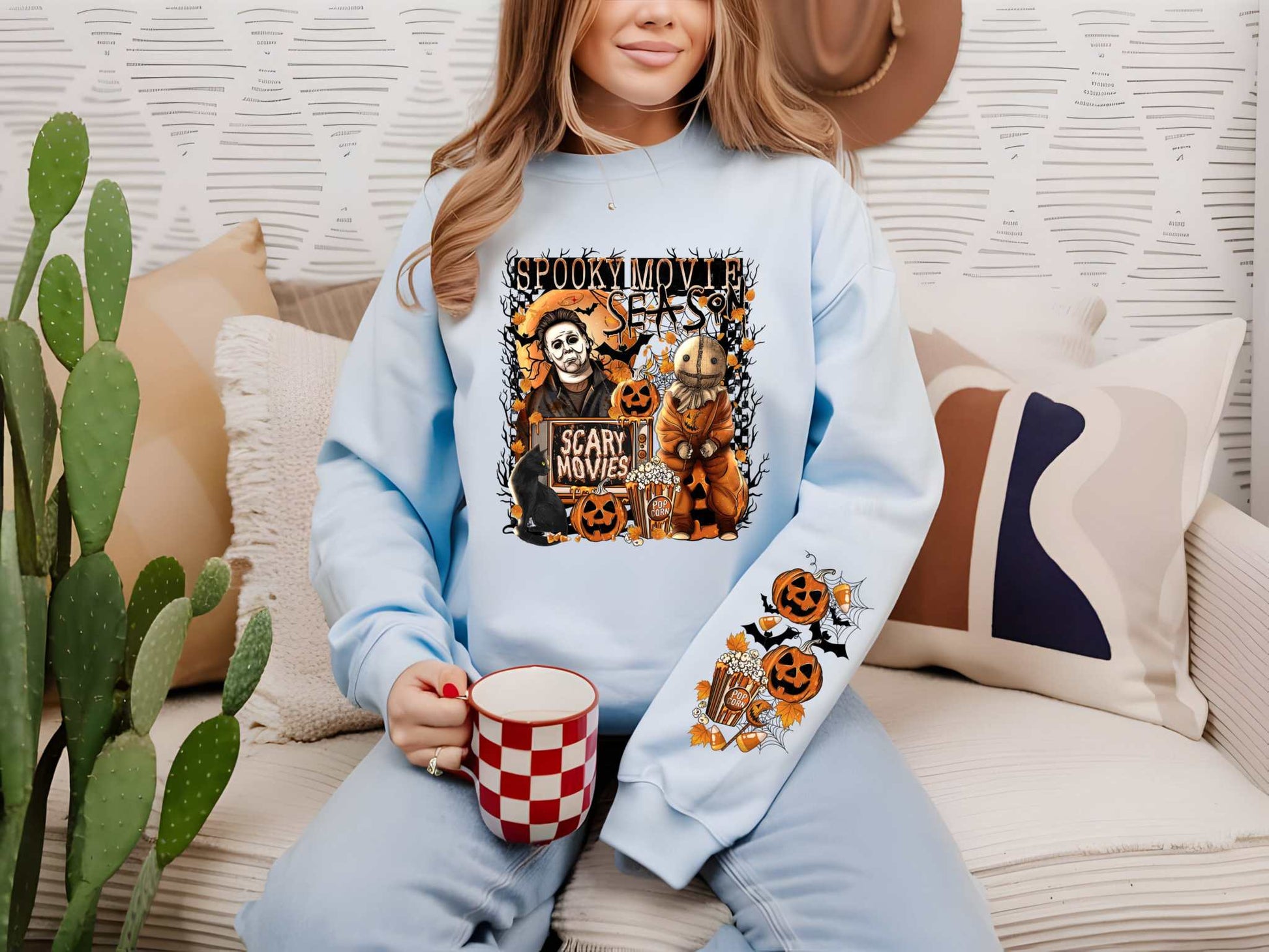 Woman wearing a Halloween-themed sweatshirt with Scary Movie Season DTF transfer, holding a checkered cup on a cozy white sofa.